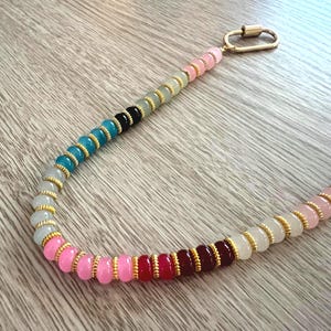 Peut inclure: Un collier de perles multicolores avec un fermoir doré. Le collier présente un dégradé de perles, passant du rose au rouge, noir, turquoise et gris, avec des accents dorés. Les perles mesurent environ 0,5 cm de diamètre.
