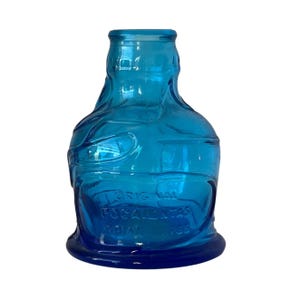 May include: A translucent blue glass bottle with a rounded body and a narrow neck. The surface features embossed text that reads THE ORIGINAL POCAHONTAS INDIAN HERBS, with a textured, organic design moulded into the glass exterior.