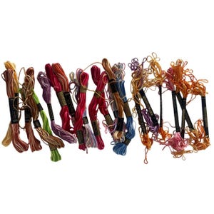 May include: A collection of multi-colored embroidery floss skeins arranged in a row. Each skein is secured with a black paper label featuring white text. The threads display a vibrant spectrum including shades of yellow, orange, red, purple, blue, and green.