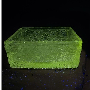 May include: A square, translucent, neon yellow glass bowl featuring an intricate embossed pattern of flowers and leaves. The glass surface displays a textured, lace-like grid design across the exterior walls, glowing brightly against a dark background.