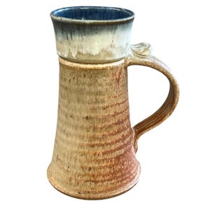 Peut inclure: Une grande tasse en céramique avec un bord évasé, présentant un corps texturé brun clair et brun rougeâtre avec des stries verticales. L'intérieur et le bord supérieur sont émaillés d'un bleu profond avec un motif de coulures blanc crème.