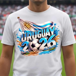 K&ouml;nnte beinhalten: Wei&szlig;es T-Shirt mit einem Grafikdesign f&uuml;r Uruguay 2026. Das Design zeigt die uruguayische Flagge, das Wort "URUGUAY" in Blau, die Zahlen "26" und einen Fu&szlig;ball. Das Design ist von blauen und goldenen Spritzern umgeben.