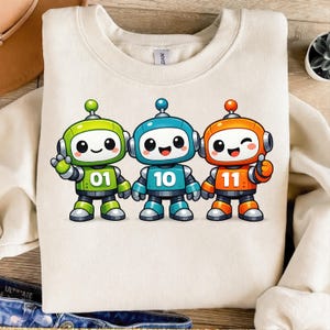 Könnte beinhalten: Cremefarbenes Sweatshirt mit drei Cartoon-Robotern in Grün, Blau und Orange. Jeder Roboter hat eine Nummer auf der Brust: 01, 10 und 11. Die Roboter haben lächelnde Gesichter und sind in einer freundlichen Pose.