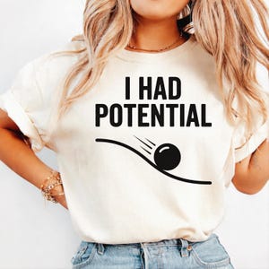 Pode incluir: Camiseta creme com o texto "I HAD POTENTIAL" em letras pretas em negrito. Abaixo do texto, um gráfico de uma bola preta rolando por uma linha curva com linhas de velocidade. A camiseta é de estilo casual.