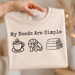 May include: Beige crew neck t-shirt with the text "My Needs Are Simple" and illustrations of a coffee cup, basketball, football, and books. The shirt is held up by a hand.
