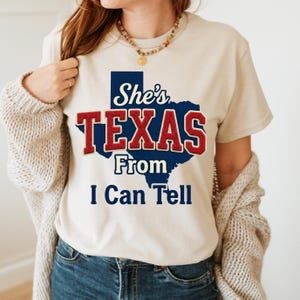 May include: Beige t-shirt with the text "She's TEXAS From I Can Tell" in a blue and red design. The word "TEXAS" is in large, bold letters, and the state outline is in blue. The shirt is a casual style.