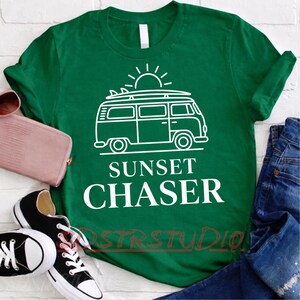 Peut inclure: T-shirt vert avec un dessin blanc d'un van surmonté d'une planche de surf, d'un soleil et l'inscription "Sunset Chaser". L'image comprend un portefeuille rose, des baskets noires et un jean bleu.