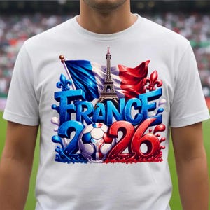 K&ouml;nnte beinhalten: Wei&szlig;es T-Shirt mit einem Grafikdesign, das Frankreich feiert. Das Design umfasst das Wort "FRANCE" in Blau, den Eiffelturm, einen Fu&szlig;ball und die Zahlen "2026" in Rot. Die franz&ouml;sische Flagge und Fleur-de-Lis sind ebenfalls abgebildet.