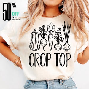 May include: Cream-colored crop top with a black graphic of vegetables including a pumpkin, beets, carrots, and onions. The words "CROP TOP" are printed in black below the vegetables. The top also has a 50% off sale graphic.