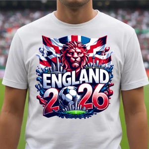 K&ouml;nnte beinhalten: Wei&szlig;es T-Shirt mit einem grafischen Design. Das Design zeigt den Text "ENGLAND 2026", einen L&ouml;wenkopf, die Flagge des Union Jack, einen Fu&szlig;ball und eine Stadtlandschaft mit roten und blauen Akzenten.