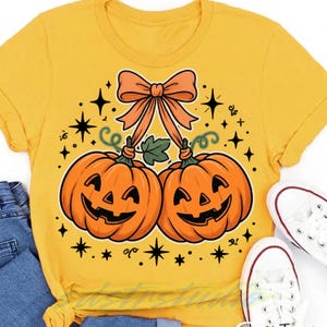 Peut inclure: Un t-shirt jaune moutarde avec un motif représentant deux citrouilles d'Halloween orange attachées par un grand nœud orange. Le design comprend des tiges de citrouille vertes, de petites vrilles vertes et des étoiles noires tout autour.