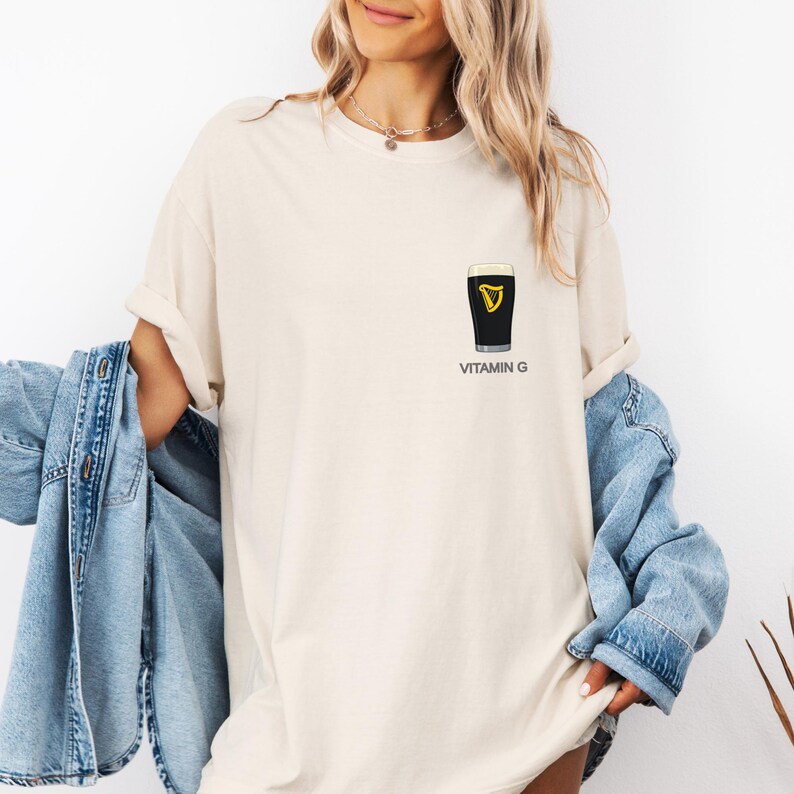 A woman wearing a white t-shirt with a graphic of a glass of Guinness beer and the text "VITAMIN G" printed on it, along with a denim jacket.