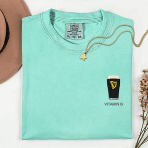 A turquoise t-shirt with a graphic of a pint of Guinness beer and the text "VITAMIN G" printed on it, along with a star-shaped charm and a gold chain necklace.