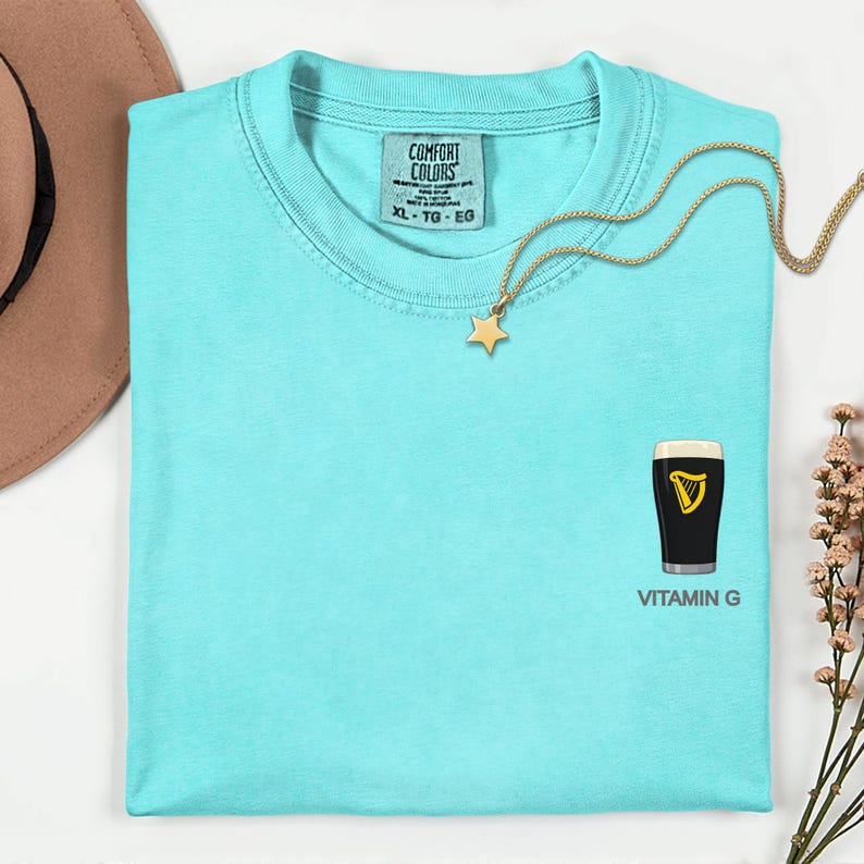A turquoise t-shirt with a vitamin G graphic and a star pendant, along with a necklace and a hat, are displayed on a white surface.
