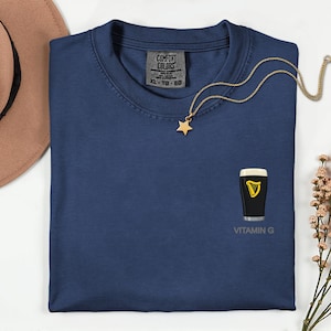 A blue t-shirt with a vitamin G graphic and a star pendant, along with a necklace and a hat, are displayed on a white surface.