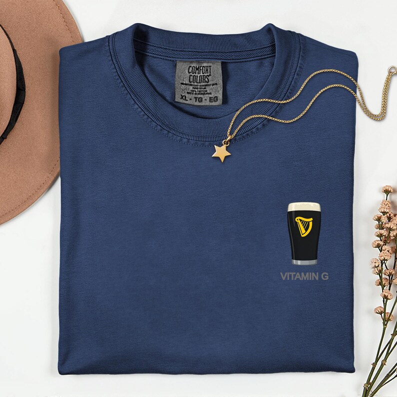 A blue t-shirt with a vitamin G graphic and a star pendant, along with a necklace and a hat, are displayed on a white surface.