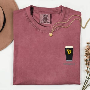 A pink t-shirt with a graphic of a pint of Guinness beer and a star-shaped charm, along with a necklace and a hat, are displayed on a white surface.