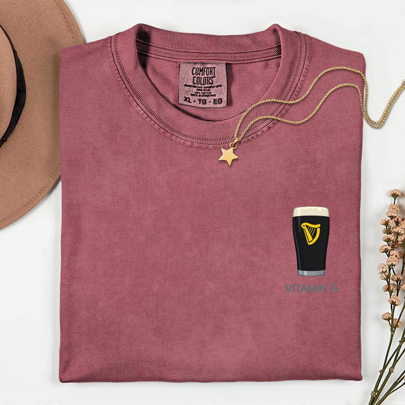 A pink t-shirt with a graphic of a pint of Guinness beer and a star-shaped charm, along with a necklace and a hat, are displayed on a white surface.