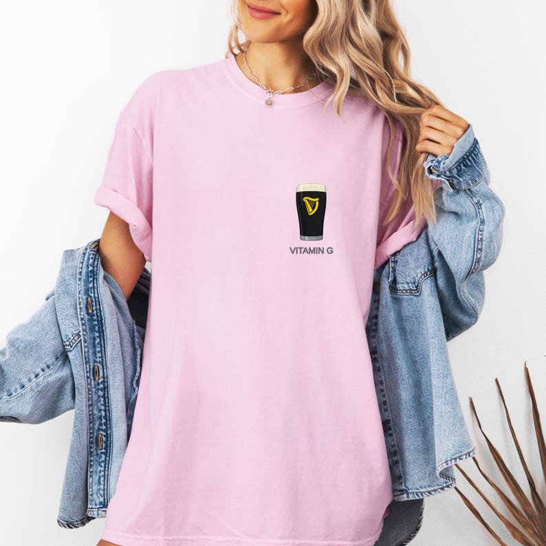 A woman wearing a pink t-shirt with a graphic of a glass of Guinness beer and the text "VITAMIN G" printed on it, along with a denim jacket.