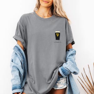 A woman wearing a gray t-shirt with a graphic of a pint of Guinness beer and the text "VITAMIN G" printed on it, along with denim clothing and a denim jacket.