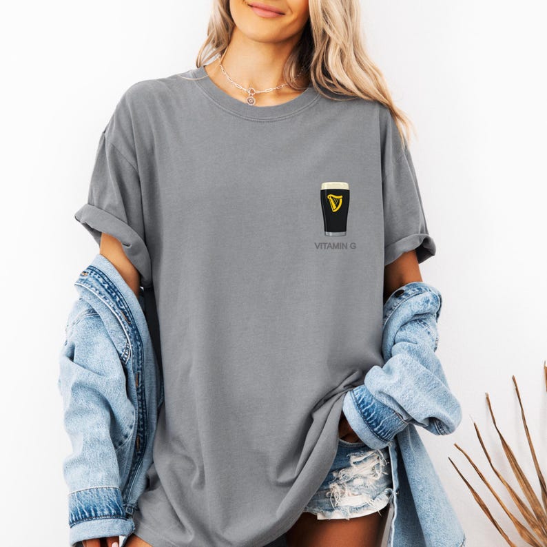 A woman wearing a gray t-shirt with a graphic of a pint of Guinness beer and the text "VITAMIN G" printed on it, along with denim clothing and a denim jacket.