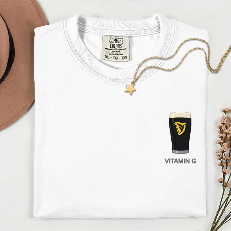A white t-shirt with a graphic of a pint of Guinness beer and the text "VITAMIN G" printed on it, along with a gold star charm and a brown hat.