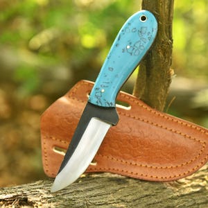 Peut inclure: Un couteau à lame fixe avec un manche turquoise vif orné d'un motif tacheté sombre et d'une goupille métallique. La lame présente une finition sombre près du dos et un tranchant poli et réfléchissant. Il est accompagné d'un étui en cuir tanné.