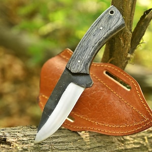 Peut inclure: Un couteau à lame fixe avec un manche texturé sombre et une lame en acier poli. Le couteau présente une mitre prononcée et un trou pour dragonne à l'extrémité du manche. Il est placé devant un étui en cuir beige avec des coutures visibles.