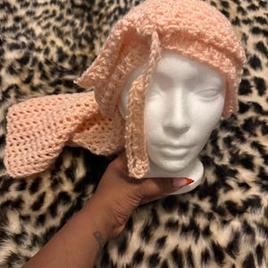 May include: A light peach-colored crochet hat and scarf set. The hat is a close-fitting style with a brim, and the scarf is long and rectangular. The set is displayed on a white mannequin head against a leopard print background.