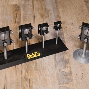May include: A display of black, silver, and grey mechanical objects, some mounted on a black base with the text "RobCo". The objects have a boxy design with silver accents and are displayed on a wooden surface.