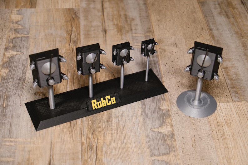 May include: A display of black, silver, and grey mechanical objects, some mounted on a black base with the text "RobCo". The objects have a boxy design with silver accents and are displayed on a wooden surface.
