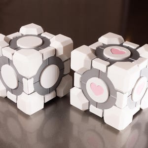 Puede incluir: Dos objetos cúbicos blancos y grises con un diseño geométrico segmentado. Cada cara presenta un centro circular. Un cubo tiene círculos blancos lisos, mientras que el otro cubo muestra un icono de corazón rosa claro centrado en cada cara.