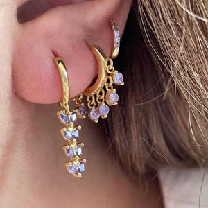 May include: Gold-coloured hoop earrings with purple gemstone embellishments. One earring has a cascade of purple gemstones, another a crescent shape with dangling gems. A smaller hoop earring with purple gems is also shown.