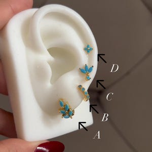 May include: A display of four different gold-colored earrings with turquoise-colored stones. The earrings are arranged on a white ear-shaped display. The earrings include a small stud, a small hoop, and two other designs.