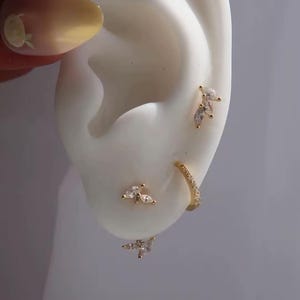 May include: A selection of gold-coloured earrings with clear, sparkling stones are displayed on a white ear model. The earrings include a small hoop, a cluster of stones, and a marquise-shaped design. The ear model is held by a person with a yellow nail.