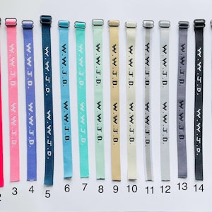 以下が含まれることがあります： 赤、ピンク、青、緑、ニュートラルカラーなど、さまざまな色の16本の時計ストラップのコレクションです。各ストラップには、白い文字で「W.W.J.D.」と書かれています。ストラップは白い背景に垂直に配置されています。