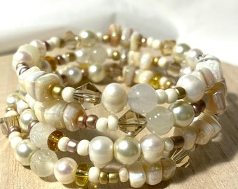 Puede incluir: Un brazalete de múltiples hilos con perlas blancas, cuentas de concha iridiscente y cuentas de vidrio doradas y transparentes.
