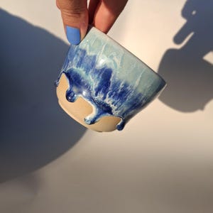 Könnte beinhalten: Eine kleine Keramiktasse mit einer matten beigen Basis und einer glänzenden Glasur in Blau- und Weißtönen, die vom Rand herabläuft. Die Glasur weist ein strukturiertes, geschichtetes Aussehen mit tiefen Marine- und hellen Himmelblautönen auf.
