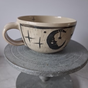 Könnte beinhalten: Cremefarbene Keramiktasse mit schwarzem Himmelsdesign. Die Tasse zeigt einen Mond mit Gesicht, Sterne und ein Kreuzmuster. Die Tasse hat einen großen Henkel und steht auf einem grauen, runden Ständer.