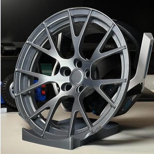 May include: A grey alloy wheel with a multi-spoke design, displayed on a matching grey stand. The wheel has a central hub and is designed for a vehicle. The background includes a computer monitor and other objects.