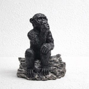 Può includere: Statuetta di scimpanzé nero seduta su un tronco strutturato di colore grigio. Lo scimpanzé è in posa pensosa, con la mano che copre la bocca. La statuetta presenta dettagli di pelliccia e tratti del viso.