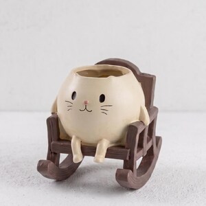May include: A cream-coloured ceramic planter shaped like a round cat head with black eyes, a pink nose, and whiskers, seated in a dark brown miniature wooden rocking chair. The cat figure has small dangling legs resting on the chair seat.
