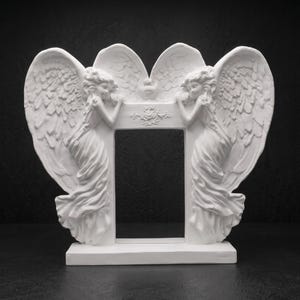 May include: A white rectangular picture frame featuring two sculpted figures with large feathered wings on either side. The figures are positioned facing inward toward the centre opening, with a small floral relief detail centred above the frame.