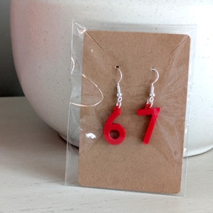 May include: A pair of red number earrings, featuring the numbers 6 and 7, displayed on a brown card within a clear plastic package. The earrings have silver-colored hooks. The background includes a white ceramic vase.