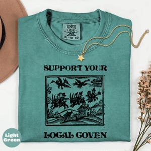May include: A teal short-sleeved t-shirt featuring a black graphic print of figures riding goats and flying creatures above houses. The text reads SUPPORT YOUR LOCAL GOVEN. A gold-toned star pendant necklace rests on the neckline of the shirt.