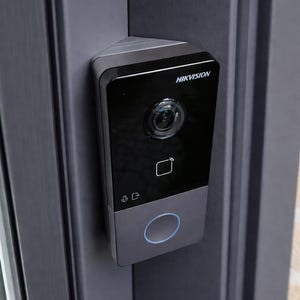 Può includere: Un videocitofono HIKVISION nero e grigio montato su un telaio di porta grigio scuro. Il videocitofono è dotato di una telecamera, un sensore touch e un anello luminoso blu. Il logo HIKVISION è visibile in alto.