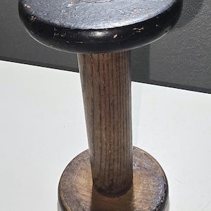 May include: A vintage wooden spool with a dark gray top and base, connected by a cylindrical, brown wooden stem. The top and base have a black trim, showing signs of wear. The spool is likely used for holding thread or yarn.