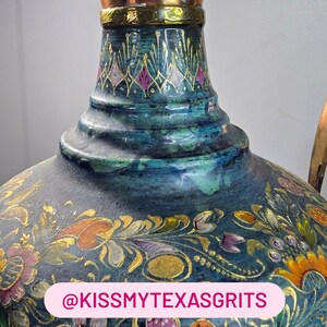 May include: A decorative ceramic vessel with a teal and blue mottled glaze, featuring intricate hand-painted floral motifs in gold, pink, and orange. The neck has a gold-toned metal band. A pink label displays the text @KISSMYTEXASGRITS.