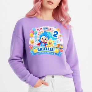 May include: A light purple long-sleeve sweatshirt featuring a colorful graphic print of the character Plim Plim, a yellow creature, balloons, a rainbow, and the text: PLIM PLIM SAY... 2 BRIELLIE! YOU ARE SO SPECIAL!