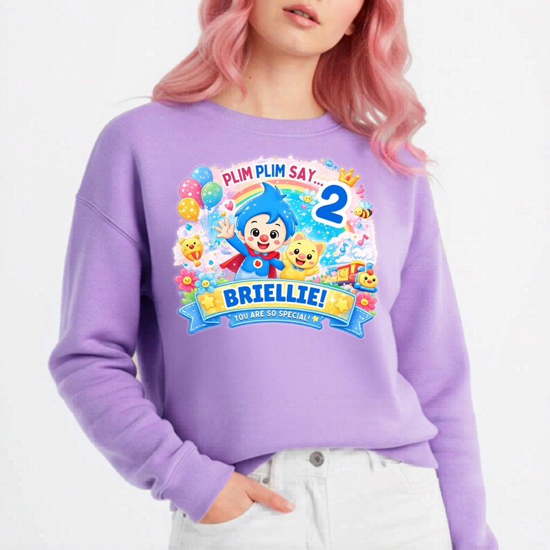 May include: A light purple long-sleeve sweatshirt featuring a colorful graphic print of the character Plim Plim, a yellow creature, balloons, a rainbow, and the text: PLIM PLIM SAY... 2 BRIELLIE! YOU ARE SO SPECIAL!